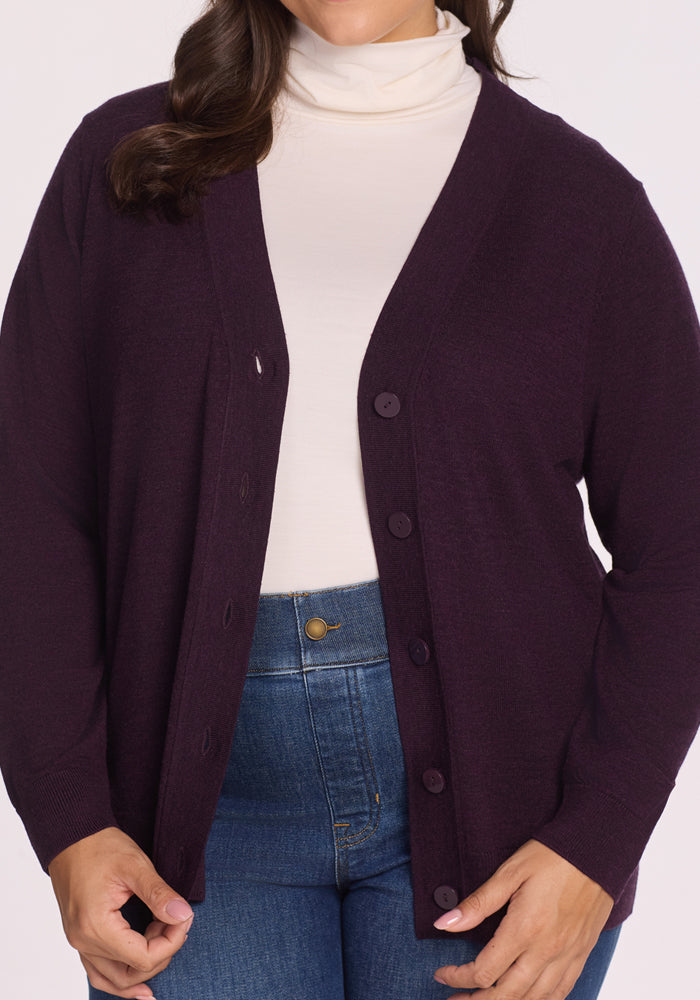 A person wearing a cream turtleneck, blue jeans, and the Woolx Gia Sweater in Deep Plum, with their head partially out of frame and hands adjusting the odor-resistant cardigan.