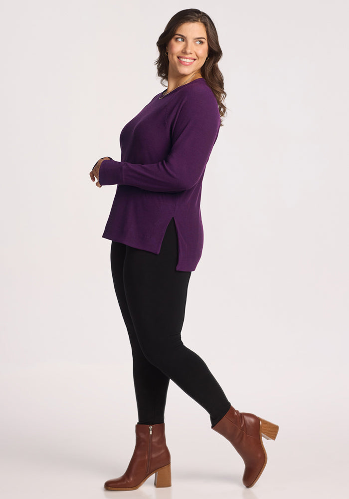 A woman with long brown hair smiles and poses sideways in the Woolx Ainsley Sweater in Majestic Purple, paired with black leggings and brown heeled ankle boots against a plain light background.