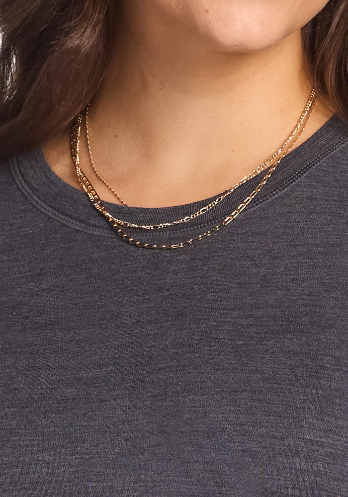 A close-up of a person wearing the Woolx Blair Tunic in Cinder Melange, paired with two thin gold chain necklaces layered around their neck. Only their lower face, neck, and part of the shoulder are visible.