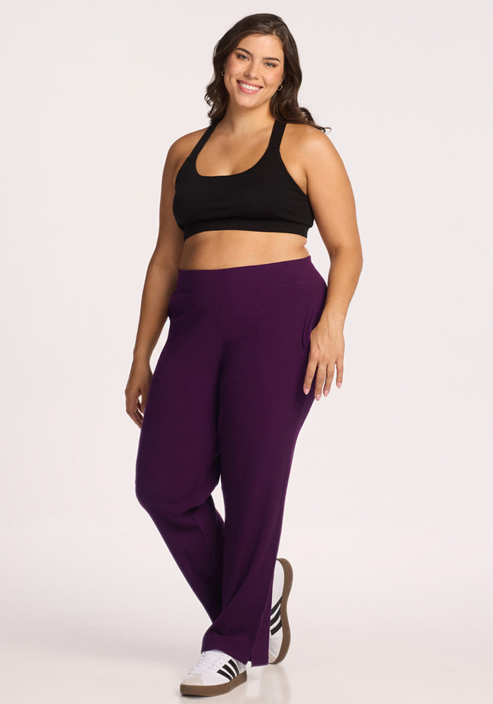 A woman smiles against a white background, wearing black sports bra, white sneakers with black stripes, and Woolx Luca Ribbed Pants in Majestic Purple.