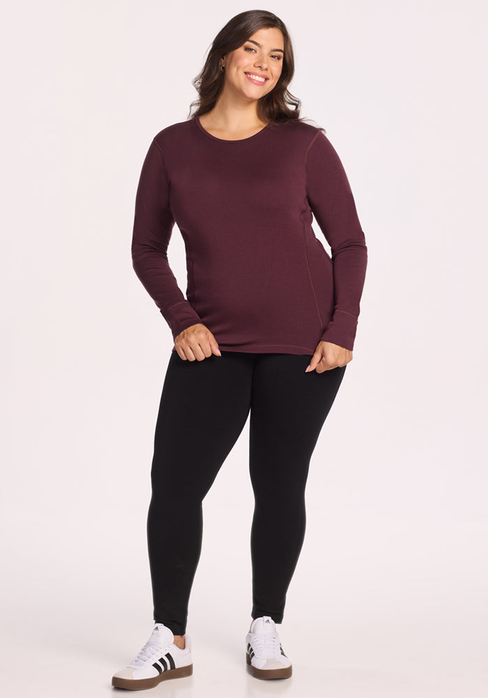 A woman smiles against a light background, wearing the Woolx Hannah Long Sleeve Top in Chocolate Fig, paired with black leggings and white sneakers with black stripes.