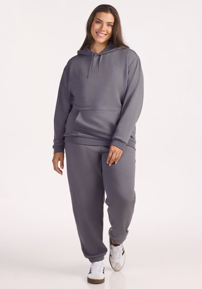 A woman smiles while wearing the Woolx Avery Hoodie in Arctic Dusk with matching sweatpants and white sneakers, radiating year-round comfort against a plain, light background.