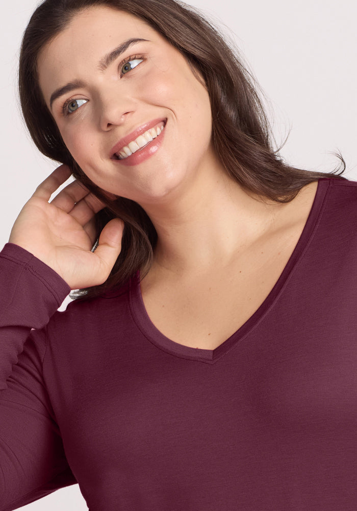 A woman with medium brown hair smiles, touching her ear. She wears the Woolx Millie Long Sleeve in Grape Harvest—a deep purple, moisture-wicking Merino wool top—while looking slightly off to the side against a plain background.