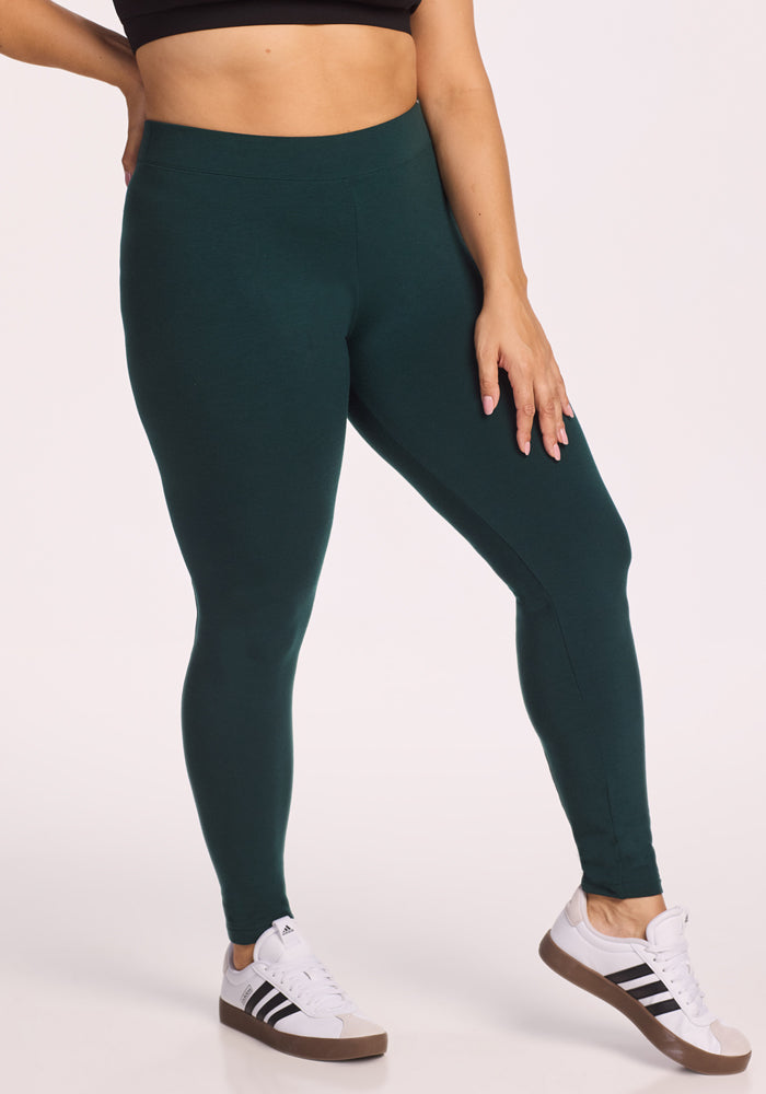 A person wearing Woolx Stella Leggings in Deep Timber, a black sports bra, and white Adidas sneakers with black stripes stands against a plain light background. Their left hand rests on their thigh.