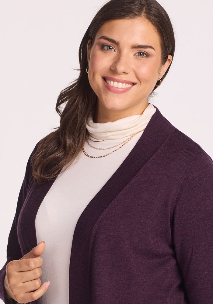A woman with long brown hair smiles warmly, wearing a white turtleneck, gold necklace, small hoop earrings, and the Woolx Macie Sweater in Deep Plum. The background is light and plain.
