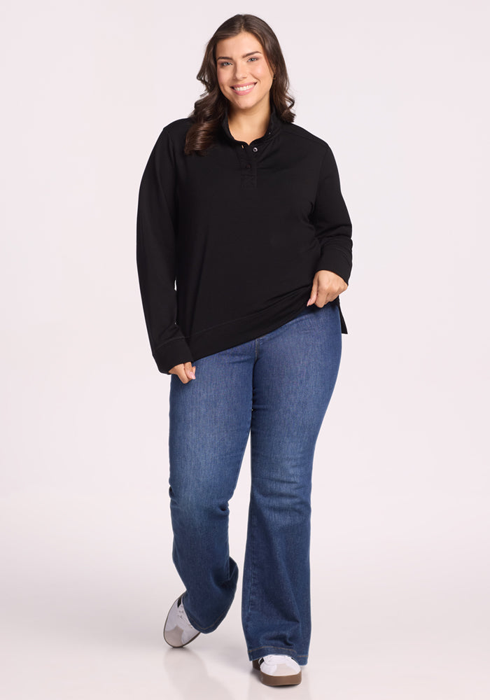 A woman with long brown hair smiles while wearing the Woolx Annie 3 Button Pullover in black, paired with blue jeans and white sneakers. She stands against a plain light background, holding the hem of her shirt with one hand.