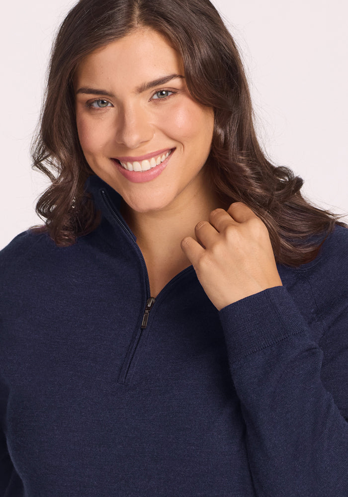 A woman with wavy brown hair and a light complexion smiles at the camera, gently holding the collar of her Woolx Collins Half Zip Sweater in Storm Cloud.