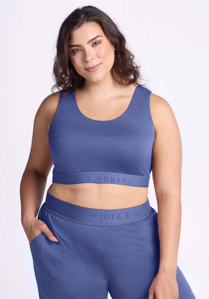 A woman with wavy brown hair smiles while wearing the Woolx Aubrey Lounge Bra in Cloudy Bay, featuring a logo band on the waistband. She stands against a light background, one hand in her pocket.