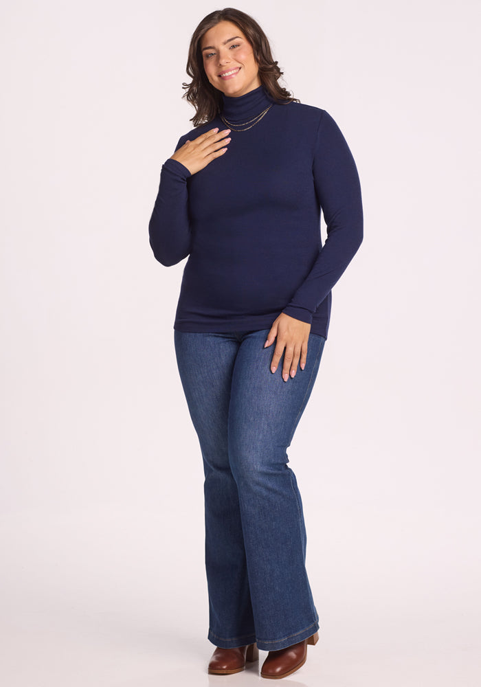 A woman with wavy brown hair wears the Woolx Sage Ribbed Turtleneck in Midnight Sapphire, blue jeans, brown ankle boots, and a necklace. She is smiling, standing, and resting one hand on her chest against a light background.