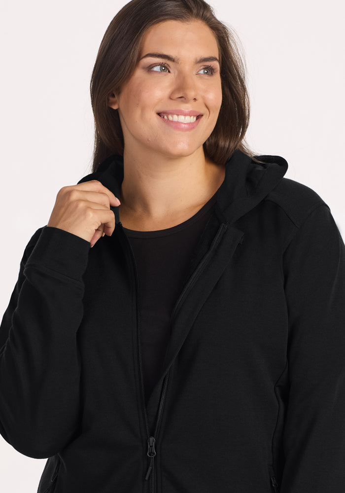 A woman with long brown hair smiles and looks to the side while holding the collar of her Woolx Darby Zip Up Hoodie in black. She wears a black shirt underneath against a plain, light-colored background.