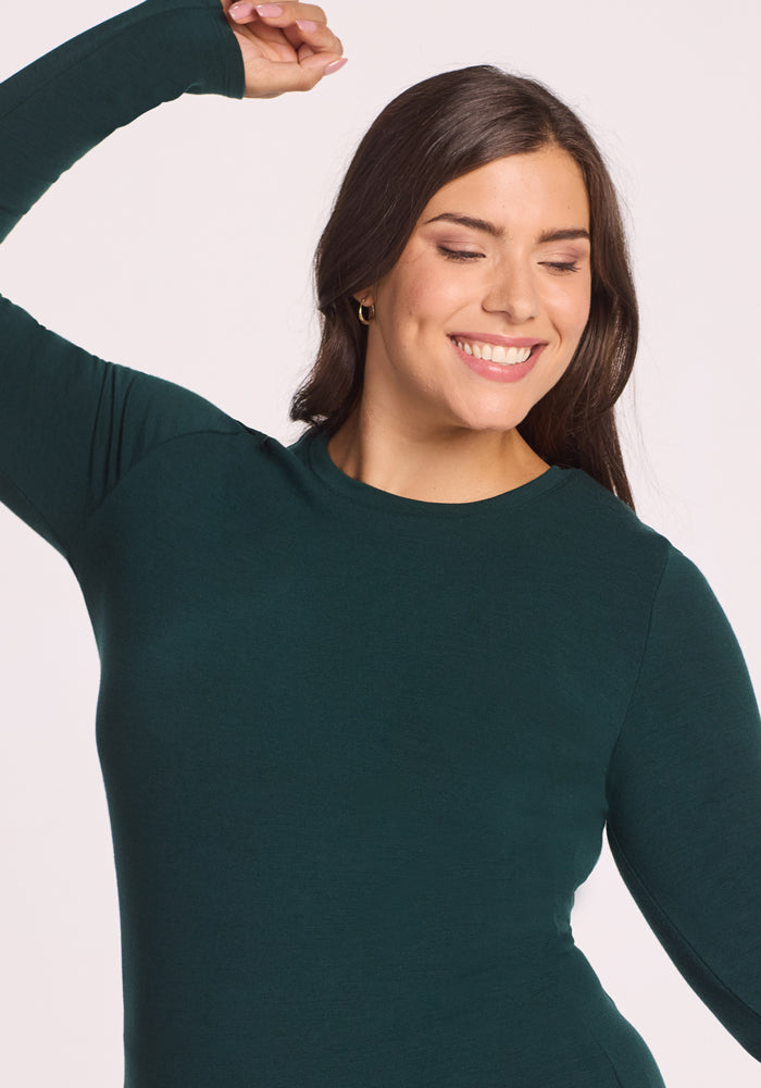 A woman with long brown hair smiles with her eyes closed, raising one arm above her head while wearing the Woolx Sadie Baselayer Top in Deep Timber, set against a light background.