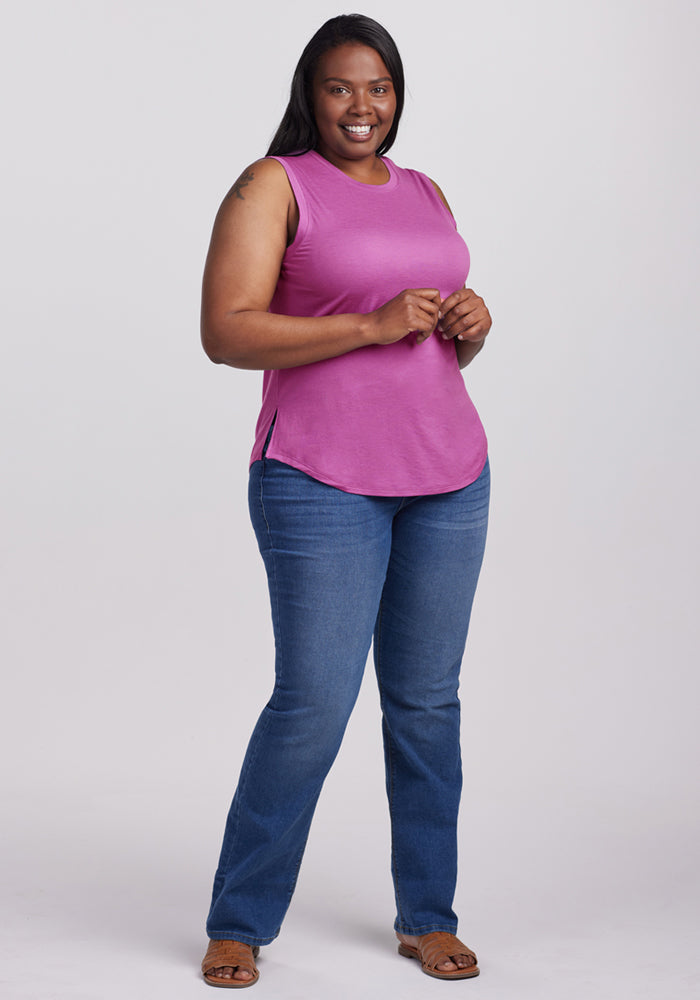 A woman with long dark hair smiles against a plain backdrop, wearing the Woolx Bella Tunic Top in Purple Wine, paired with blue jeans and brown sandals—a stylish summer look.