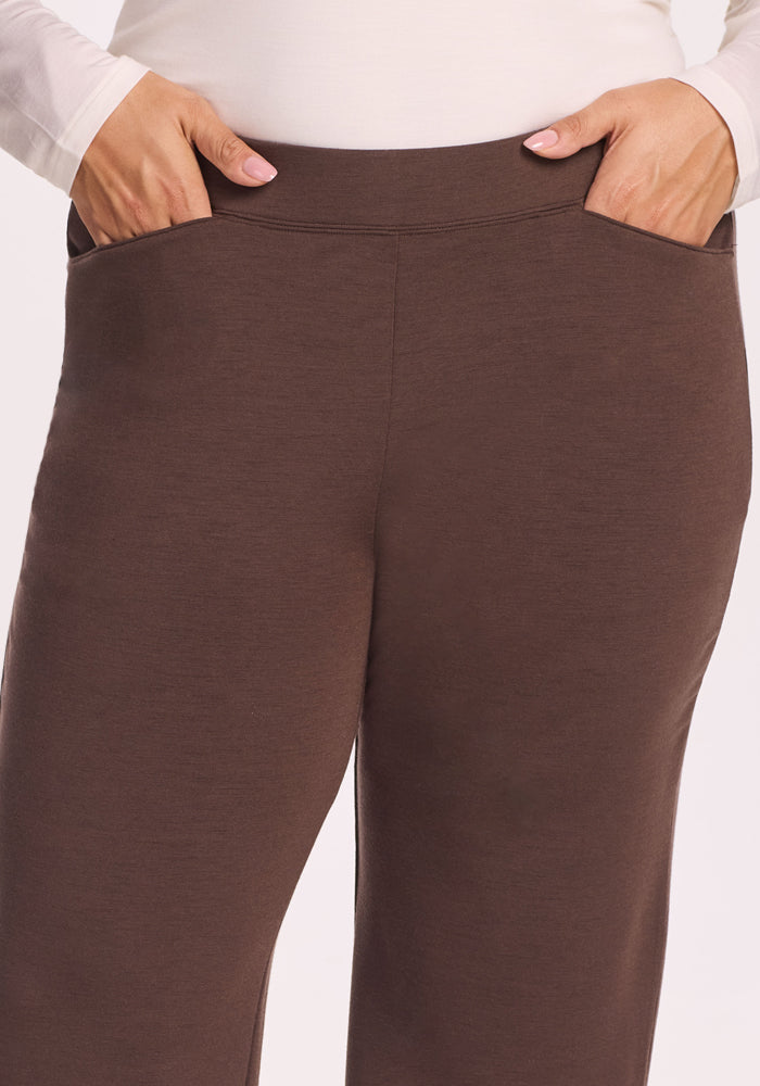 Close-up of a person wearing Woolx Ellie Wide Leg Pants in Bracken, hands in the pockets, with a white long-sleeve shirt at the waist. These warm Merino wool pants are a great thermal option. Background is plain and light-colored.