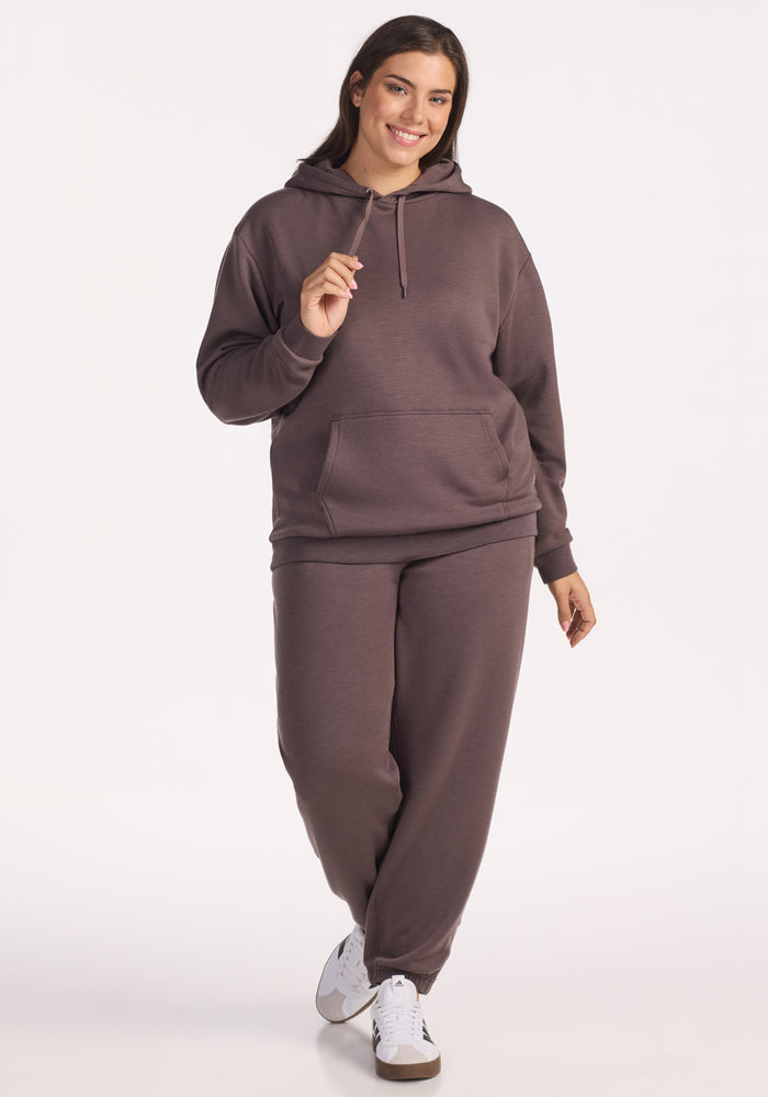 A woman smiles and walks forward wearing the Woolx Avery Hoodie in Plum Truffle with matching sweatpants and white sneakers, set against a plain white background.