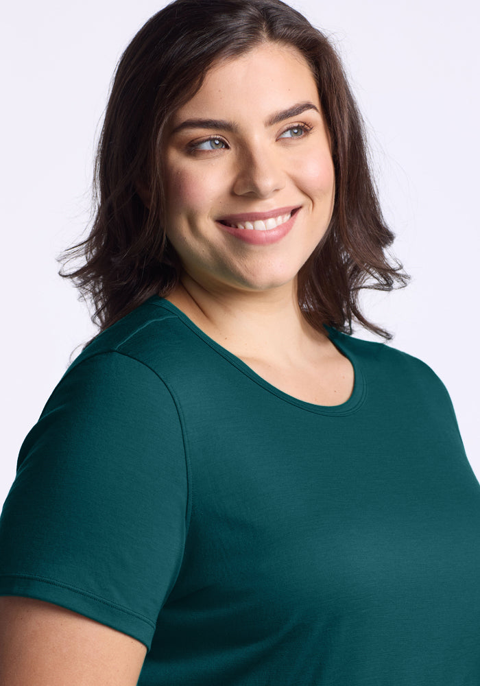 A woman with shoulder-length brown hair smiles and looks to the side, wearing the Woolx Liza Crew Neck Tee in Riverbank Green against a plain light background.