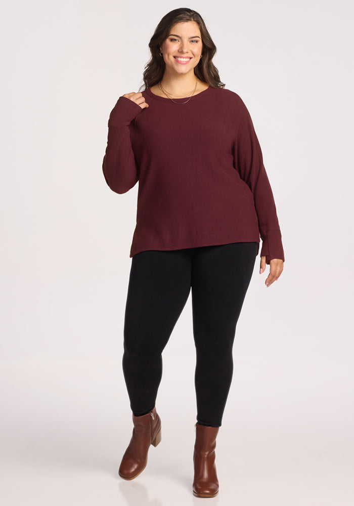 A woman with long brown hair smiles while standing and wearing the Woolx Ainsley Sweater in Toasted Garnet, paired with black leggings and brown ankle boots, against a plain light background.