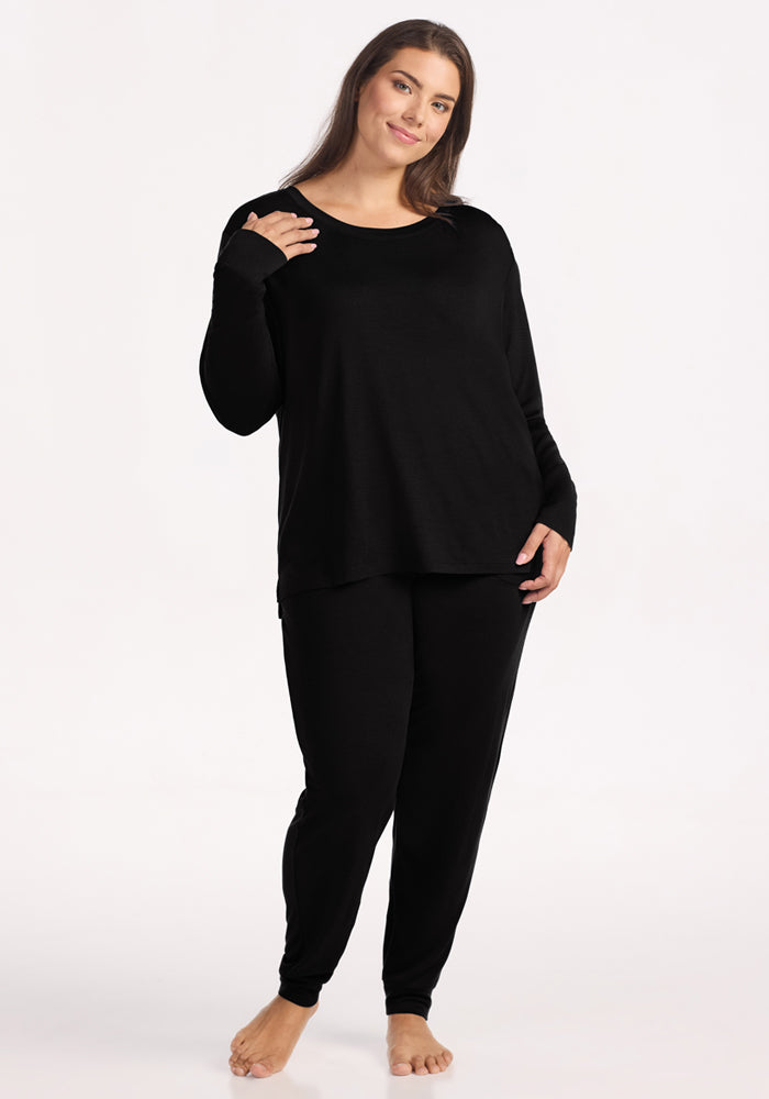 A woman stands barefoot against a light background, smiling softly with one hand on her hip. She wears the Woolx Ashlie Lounge Top in black—loose-fitting, wrinkle-resistant Merino wool—paired with matching black pants.