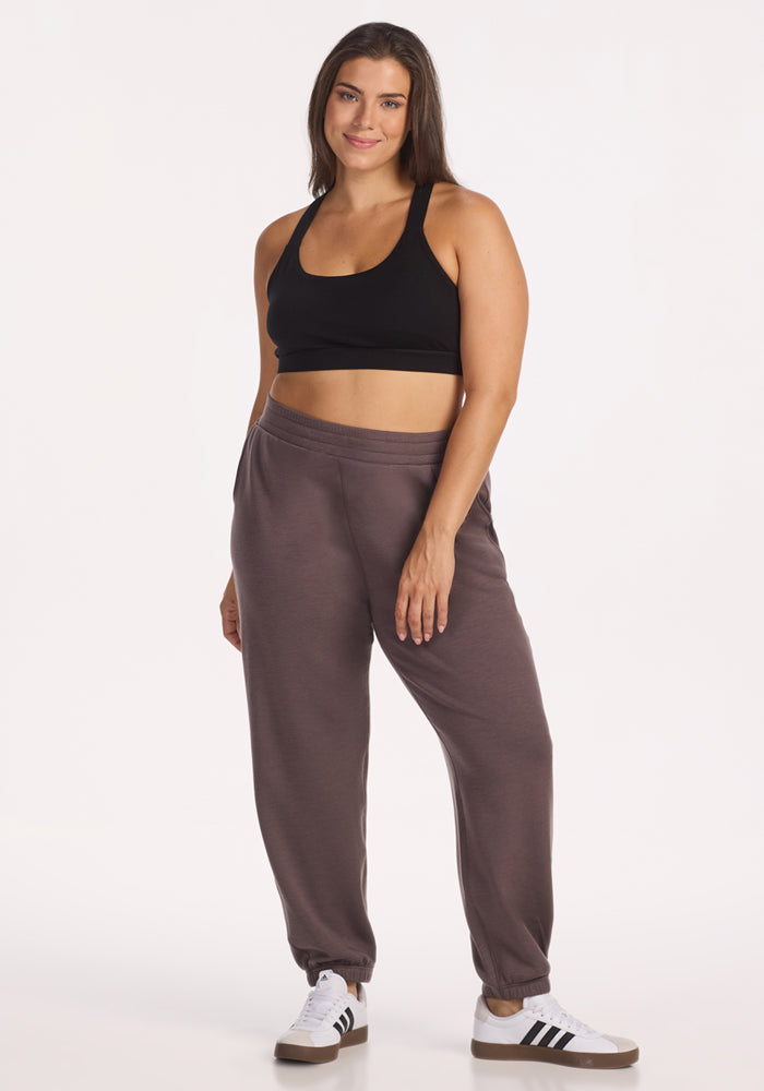 A woman stands smiling against a plain white background, wearing a black sports bra, Woolx Parker Sweatpants in Plum Truffle, and white Adidas sneakers with black stripes.