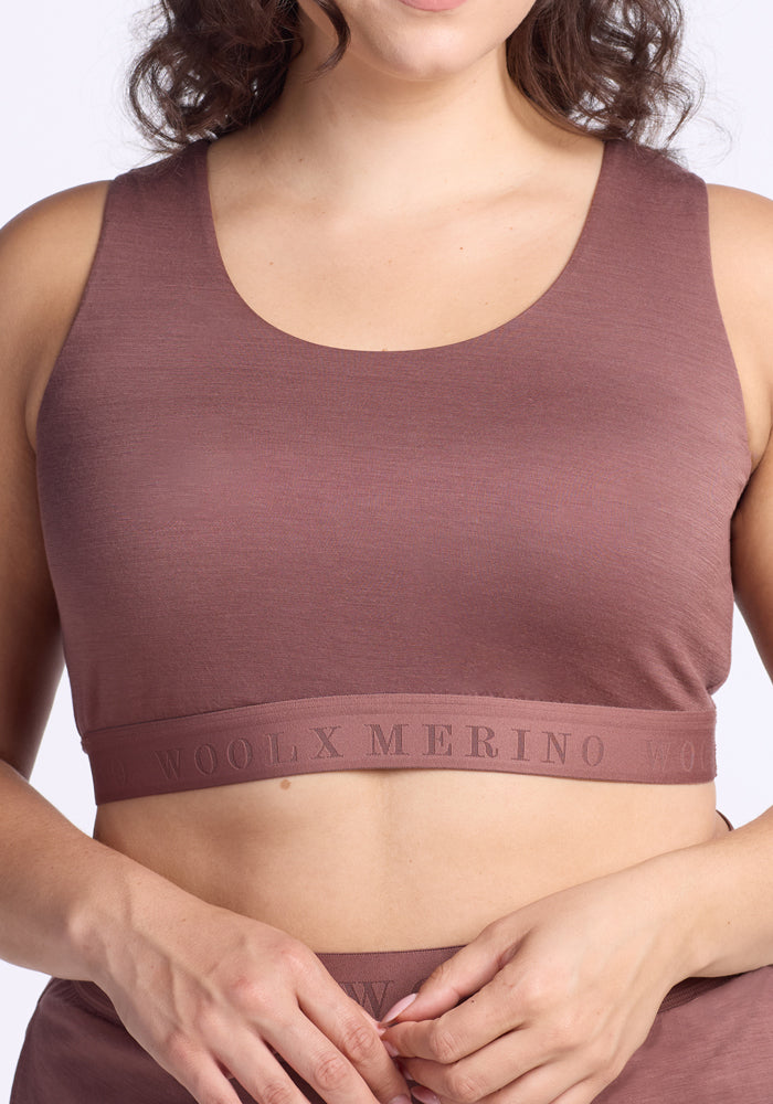 A woman wears the Woolx Aubrey Lounge Bra in Nutmeg, featuring a sleeveless design and a branded logo band, standing against a plain, light background.