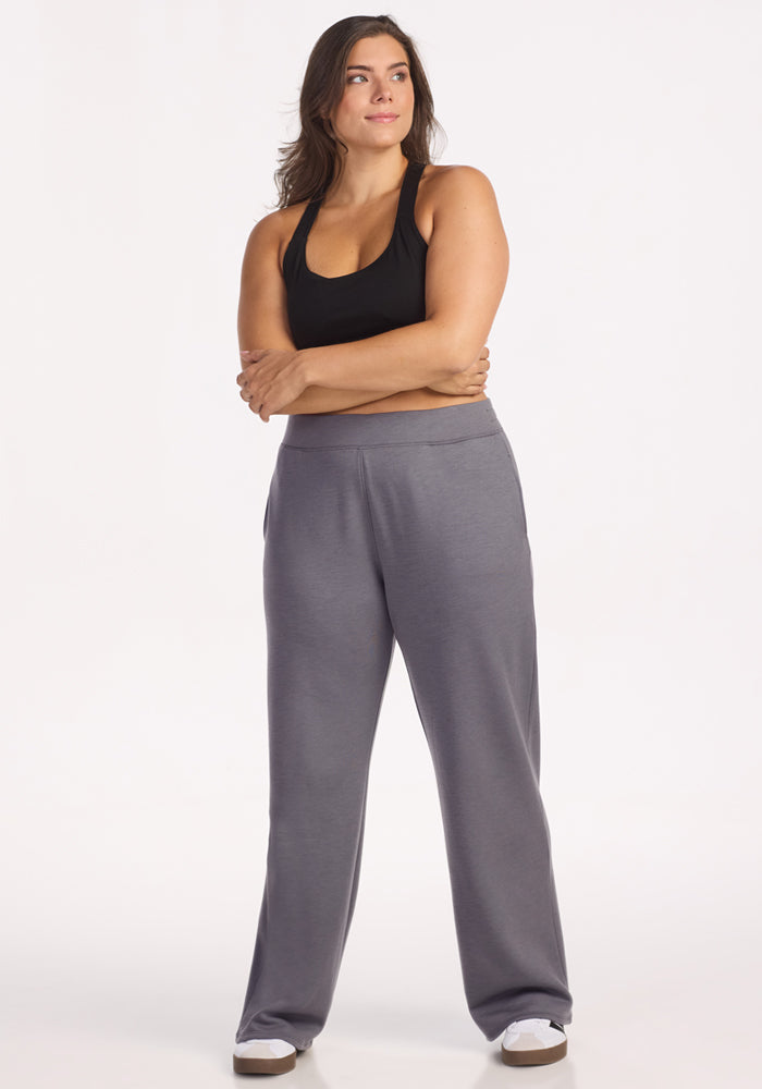 A woman stands with arms crossed, wearing a black tank top, Woolx Bree Lounge Pants in Arctic Dusk, and white sneakers. She faces slightly to the side and gazes into the distance against a plain white background.