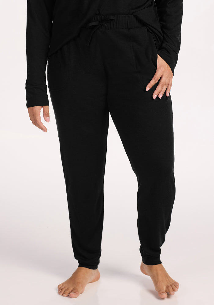 A person stands barefoot on a white background, wearing Woolx Mary Kate Lounge Pants in black and a matching black long-sleeve top with one hand in the pocket—ideal travel loungewear with soft Merino wool comfort and moisture-wicking performance. 