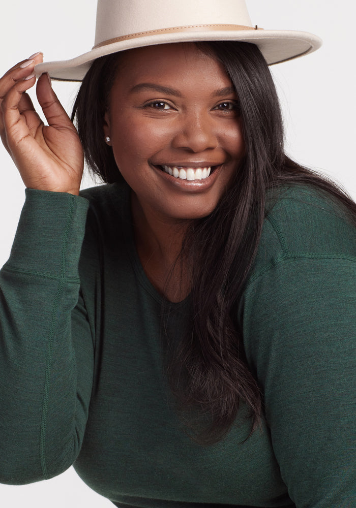 A woman with long dark hair, wearing the Woolx Hannah Long Sleeve Top in Forest and a cream-colored hat, smiles brightly at the camera while holding the brim of her hat.