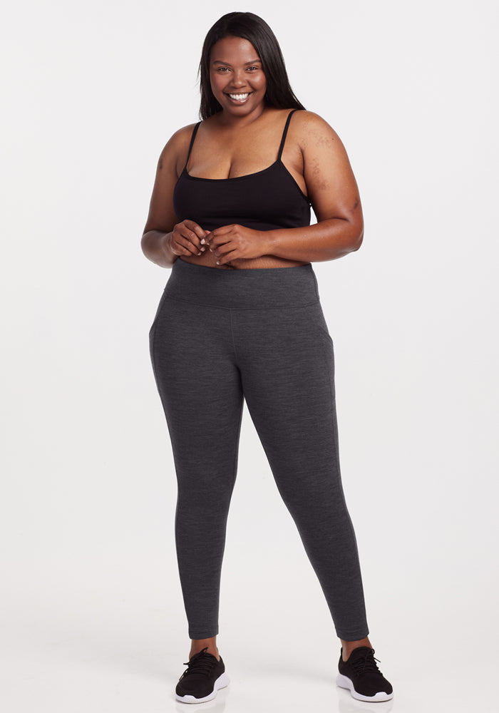 A smiling woman stands facing forward against a white background, wearing a black tank top, black sneakers, and Woolx Piper Pocket Leggings in Cinder Melange with her hands gently clasped.