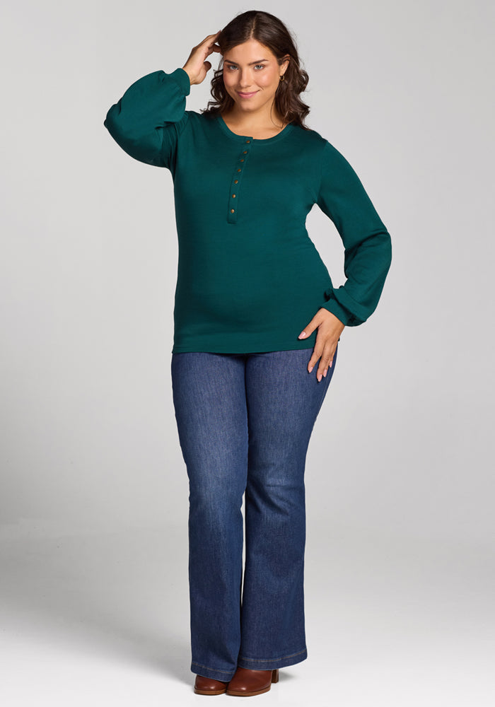 A woman with wavy brown hair wears the Woolx Charlotte Puff Sleeve Top in Riverbank Green, paired with blue bootcut jeans and brown shoes. She stands smiling, one hand touching her head, against a plain light gray background.