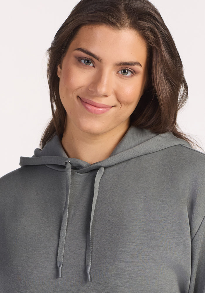 A woman with long brown hair smiles softly, wearing the Woolx Avery Hoodie in Sedona Sage, with drawstrings, against a plain light background.