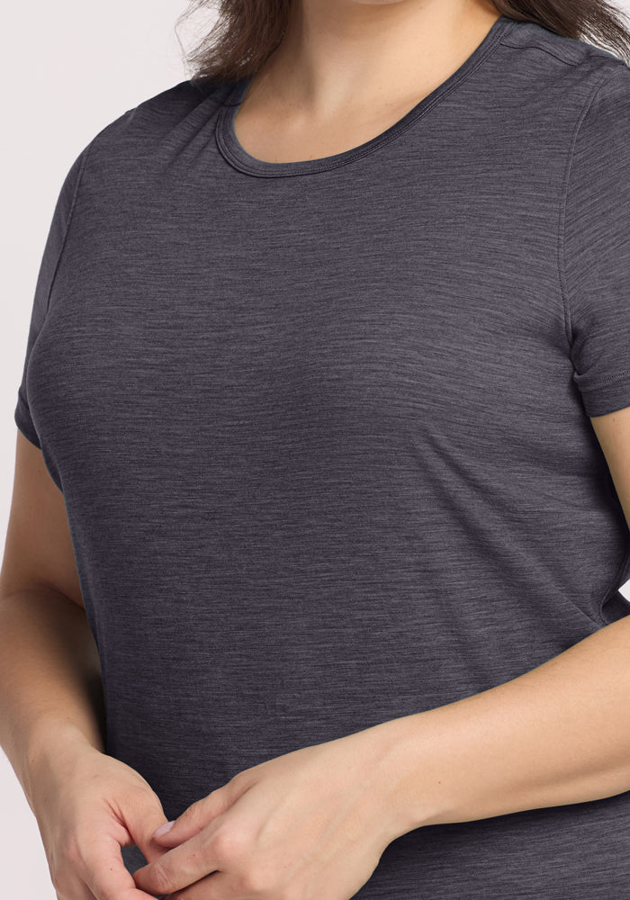 A person is shown from shoulders to waist wearing the Woolx Liza Crew Neck Tee in Cinder Melange, a short-sleeved dark gray lightweight top. Their hands are gently clasped in front against a plain, light background.