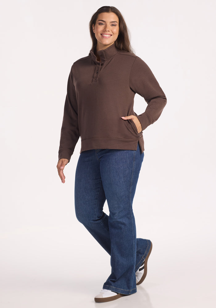 A woman smiles, wearing the Woolx Annie 3 Button Pullover in Bracken, paired with blue jeans and brown shoes. She poses with one hand in her pocket and the other relaxed by her side, standing against a plain white background.