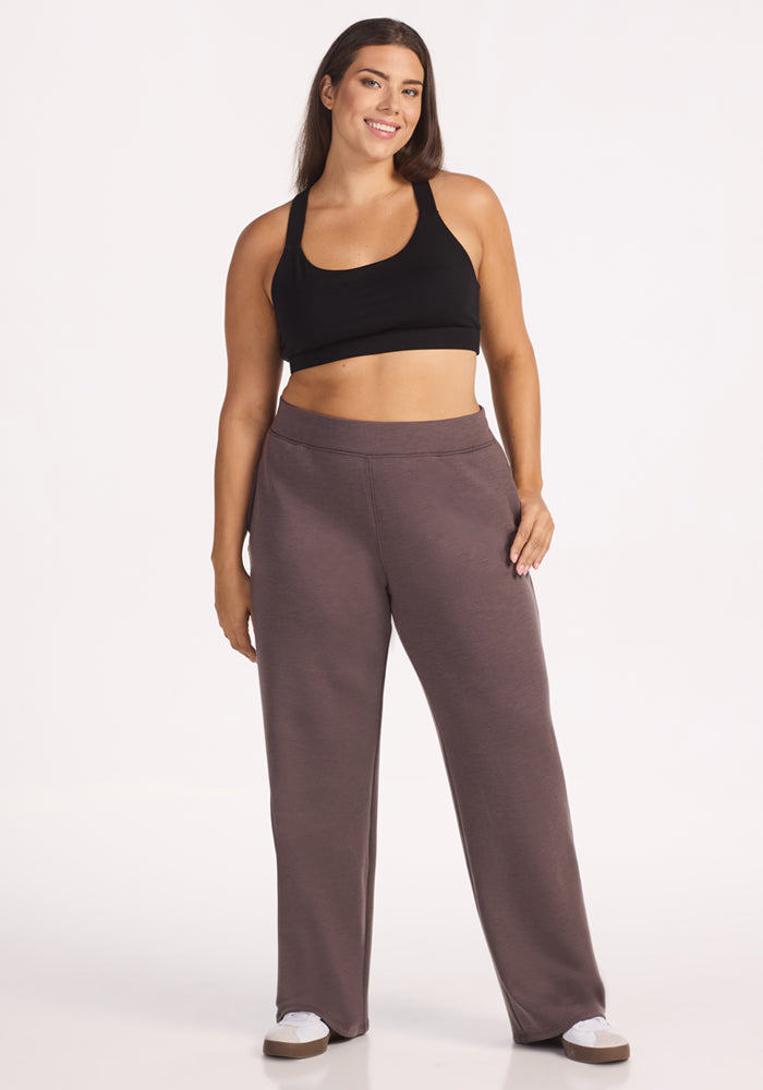 A woman with long brown hair smiles, wearing a black sports bra, Woolx Bree Lounge Pants in Plum Truffle with a relaxed straight-leg fit, and white sneakers, standing against a plain white background.