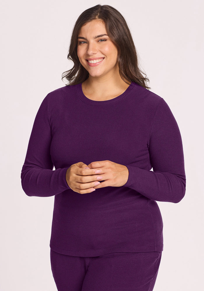 A woman with long dark hair smiles in the Woolx Hadley Ribbed Crew in Majestic Purple, made from odor-resistant, moisture-wicking fabric. She stands against a plain light background with her hands gently clasped in front of her. 