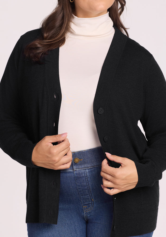 A woman is shown from shoulders to hips, adjusting the front of her unbuttoned Woolx Gia Sweater in black, worn over a white turtleneck and blue jeans.