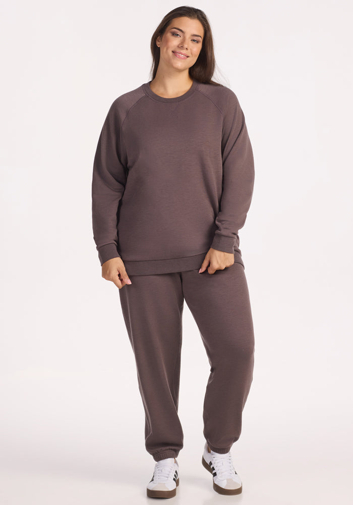 A woman smiles at the camera wearing the Woolx Bailey Crewneck Sweatshirt in Plum Truffle with matching sweatpants and white sneakers, standing against a plain white background.