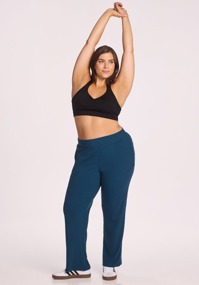 A woman wearing Woolx Luca Ribbed Pants in Majolica Blue, a black sports bra, and white sneakers stands on one leg with arms raised above her head against a plain light background.