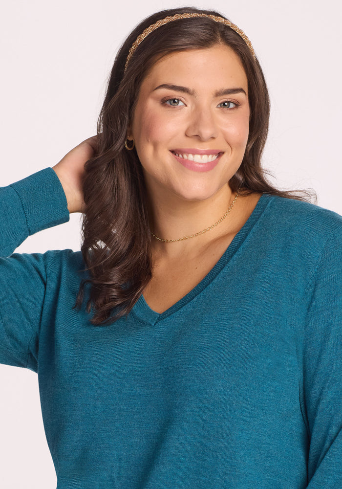 A woman with wavy brown hair, in a gold headband and jewelry, smiles at the camera. She wears the Woolx Francesca Sweater in Harbor Teal, with one hand behind her head against a plain light background.
