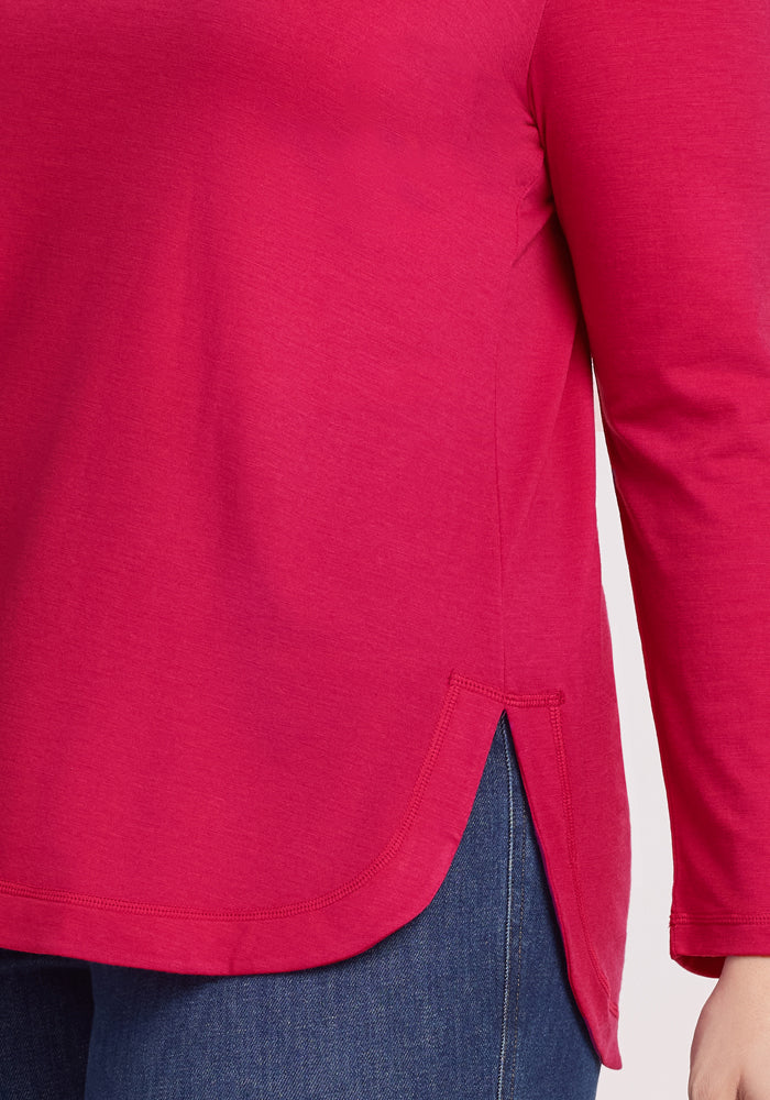 A person is shown wearing the Woolx Hazel Tunic in Poppy Spark, a long-sleeve merino wool top with a side slit hem, paired with blue jeans. Only their torso and part of one arm are visible.