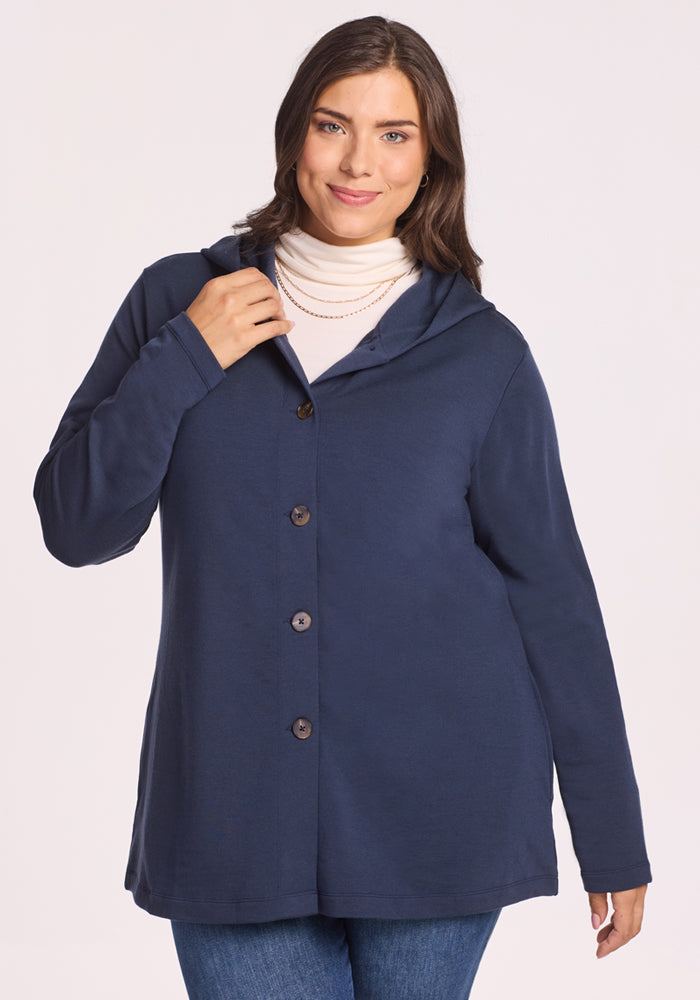 A woman with long brown hair smiles while wearing the Woolx Edna Hooded Cardigan in Deep Navy over a cream turtleneck and blue jeans, holding one side of the cardigan near the collar. 