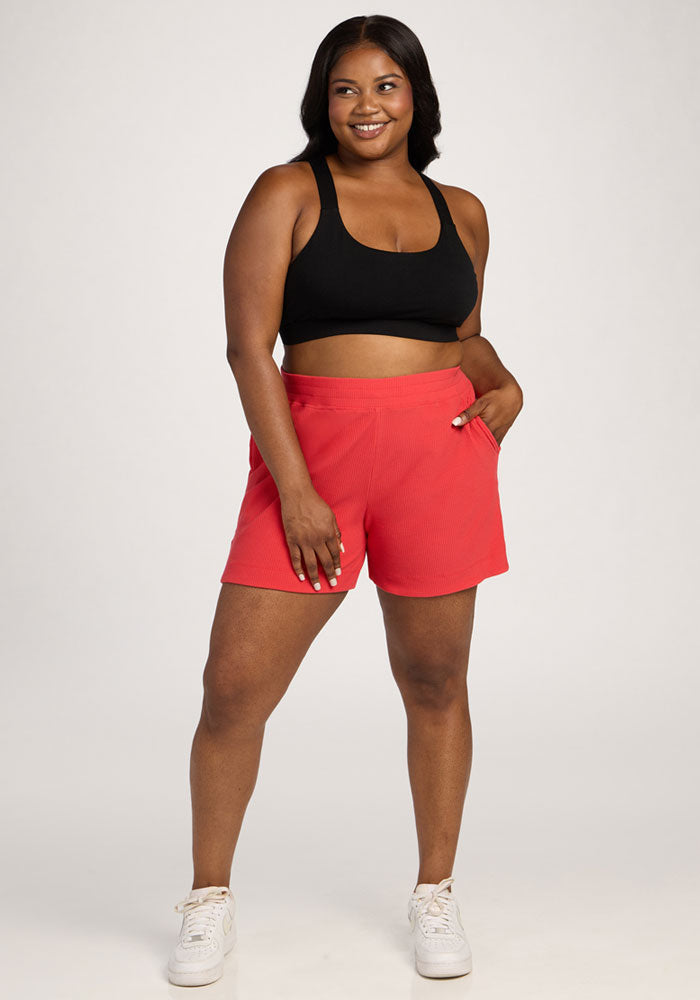 Smiling woman in a black sports bra, Woolx Maisey Ribbed Shorts - Hibiscus, and white sneakers stands hand-in-pocket. 
