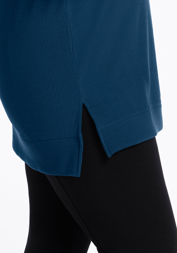 Close-up of the Woolx Evie Ribbed Tunic in Majolica Blue, showing its ribbed Merino wool texture and small side slit, styled over black leggings. The temperature regulating fabric contrasts with the white background.