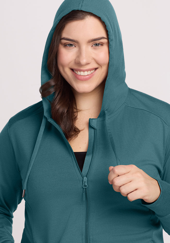 A woman with long brown hair smiles at the camera, wearing the Woolx Ryann Hoodie in Tidepool with the hood up and holding a drawstring—an essential piece for travel.