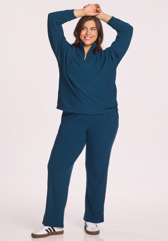 A smiling woman stands with arms raised, wearing the Woolx Quinn Ribbed 1/4 Zip in Majolica Blue and matching pants with white sneakers. The background is plain white.