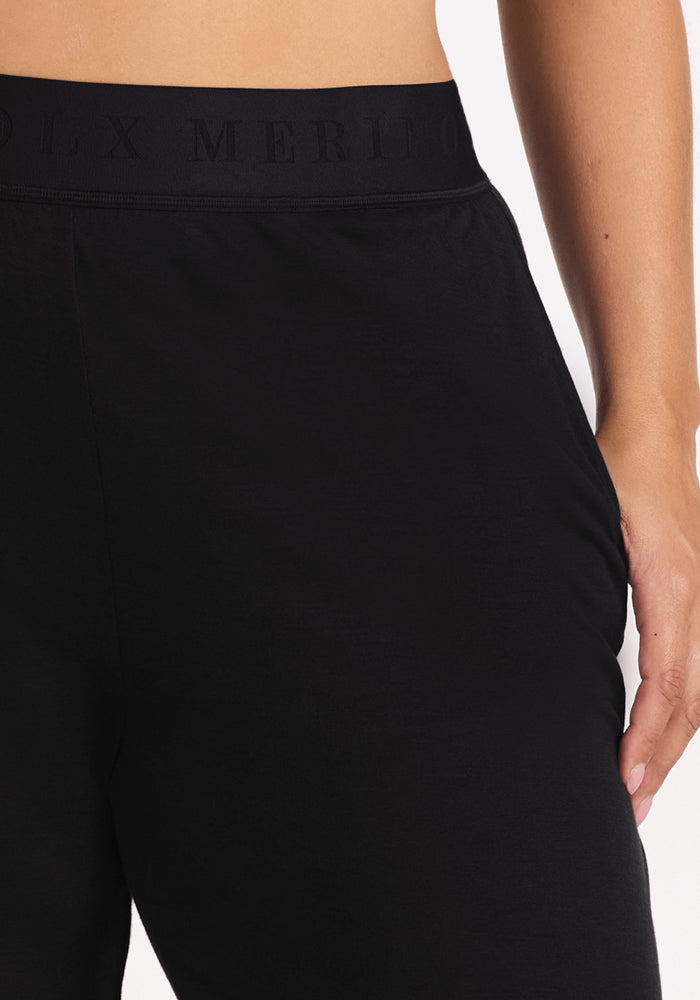 Close-up of a person wearing Woolx Maya Lounge Pants in black, featuring an elastic waistband and subtle embossed text. The temperature-regulating Merino wool pants are shown against a plain, light background with their right hand by their side.