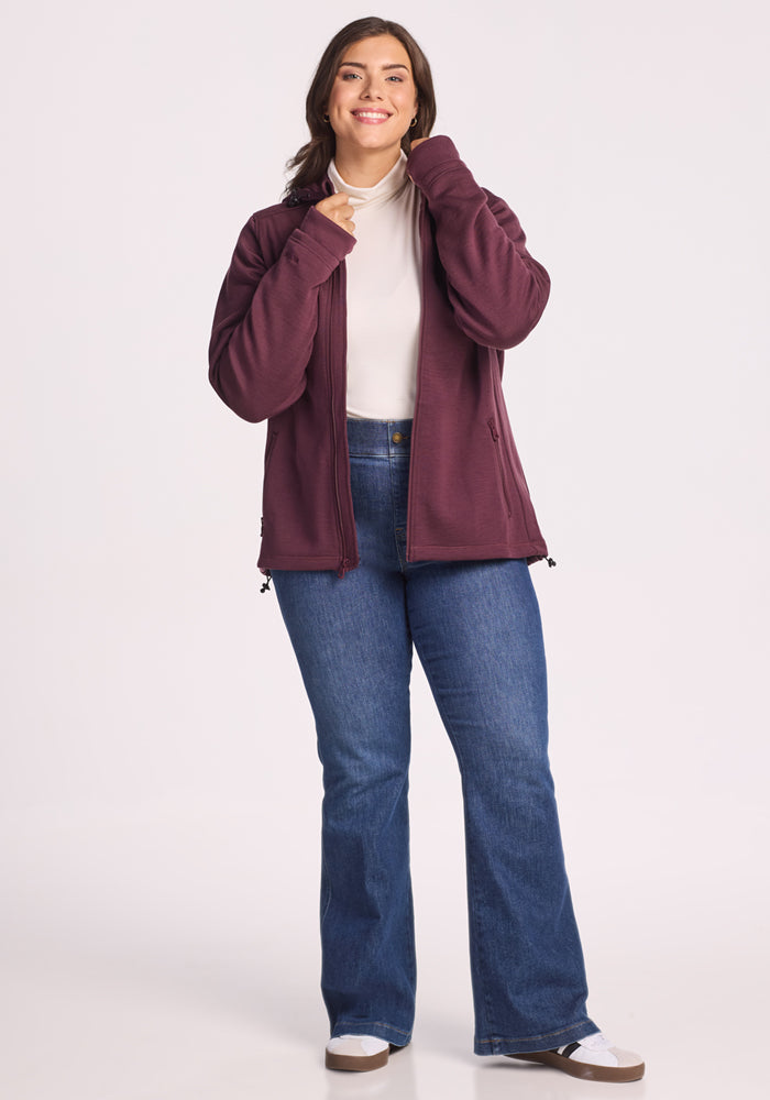 A woman smiles, wearing the Woolx Cubby Hooded Sweatshirt in Grape Harvest over a white turtleneck, with blue jeans and white sneakers, posing against a plain light background.