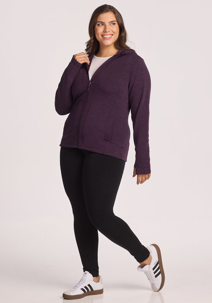 A woman smiles in casual attire: a Woolx Zoey Zip Up Hoodie in Deep Plum, black leggings, and white sneakers with black stripes, standing against a plain white background.