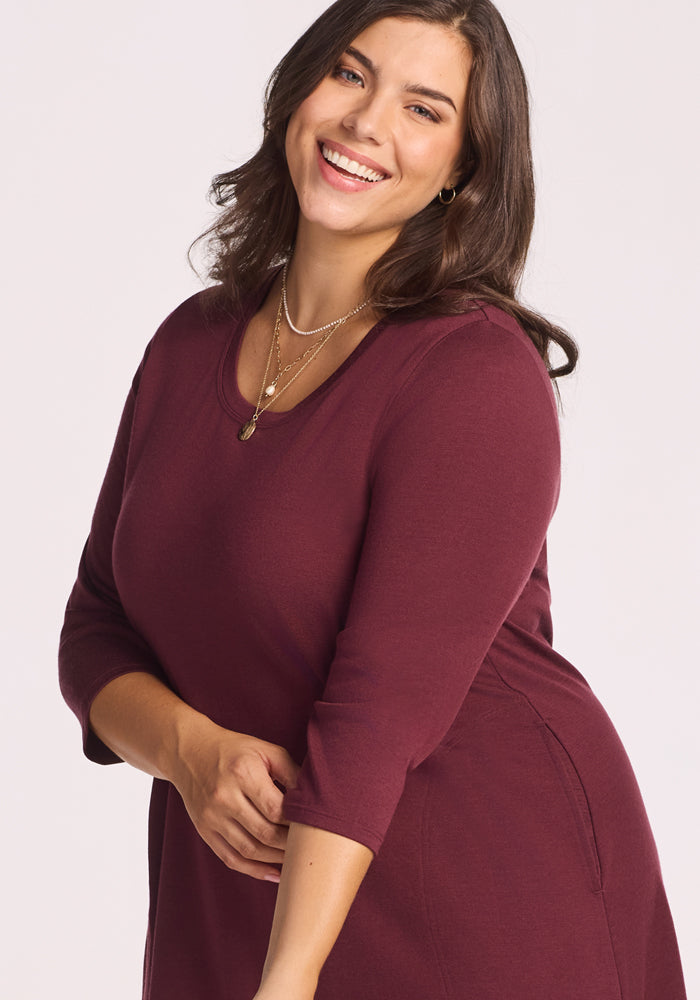 A woman with medium skin and wavy brown hair smiles at the camera, wearing the Woolx Rissa A Line Dress in Toasted Garnet, styled with layered gold necklaces against a plain light background.