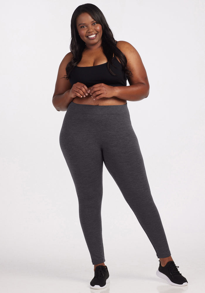 A smiling woman with long dark hair stands confidently against a white background, wearing Woolx Stella Leggings in Cinder Melange, a black crop top, and black sneakers.