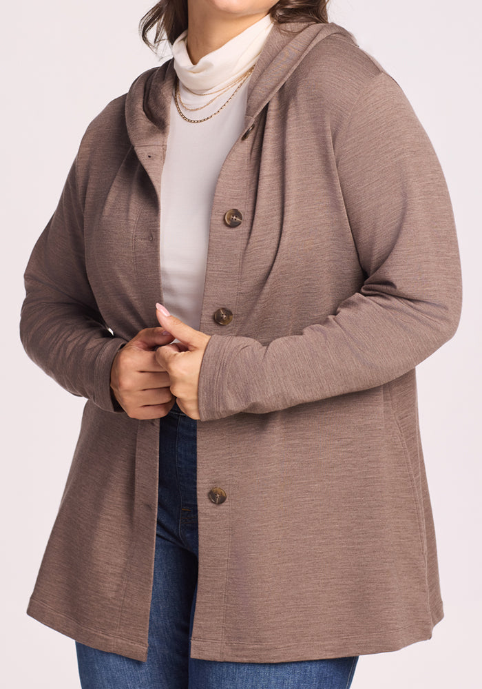 A woman styles the Woolx Edna Hooded Cardigan in Simply Taupe over a white turtleneck and blue jeans, accessorized with layered gold necklaces—an effortlessly chic cold weather look.