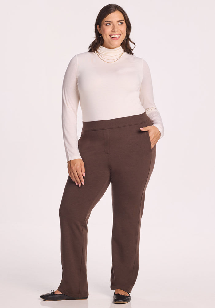 A woman smiles with one hand in her pocket, wearing a white turtleneck, black flats, a pearl necklace, and Woolx Emerson Straight Leg Pants - Bracken, posing against a plain white background.